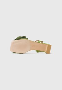 Sandalo in tessuto verde con tacco spesso e accento floreale sulla parte superiore. La suola è di un colore beige liscio con una leggera texture.