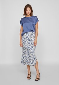 Blå satengbluse med rullede ærmer, parret med en hvid midi-nederdel med blomsterprint. Sorte stripsandaler fuldender outfittet.