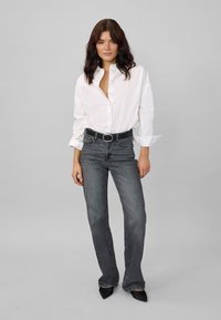 Ženska, oblečena v široko belo srajco z gumbi, črnim pasom, sivimi kavbojkami z visokim pasom in črnimi koničastimi čevlji, stoji ob sivi podlagi.