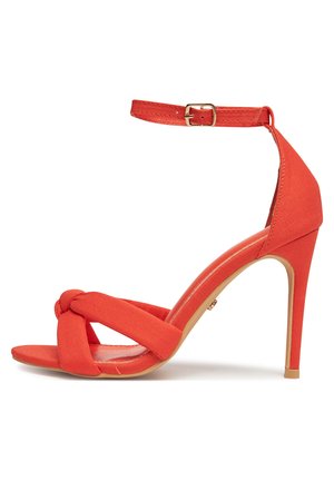 Czerwona sandałka na wysokim obcasie z wiązanymi paskami na palcach, paskiem wokół kostki z złotą klamrą oraz cienkim obcasem typu szpilka.