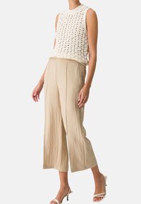 Canotta crema a maglia con design a trama aperta, abbinata a pantaloni ampi di colore beige chiaro. La modella indossa sandali bianchi con cinturini.