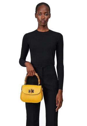 Borsa a tracolla gialla in pelle con un design strutturato, manico in stile bambù e una chiusura decorativa. Modello che indossa un top e pantaloni neri.