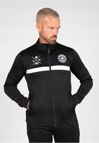 Zwarte zip-up jas van gladde stof, met een witte horizontale streep en logo's op de borst. Heeft een hoge kraag en zijzakken.