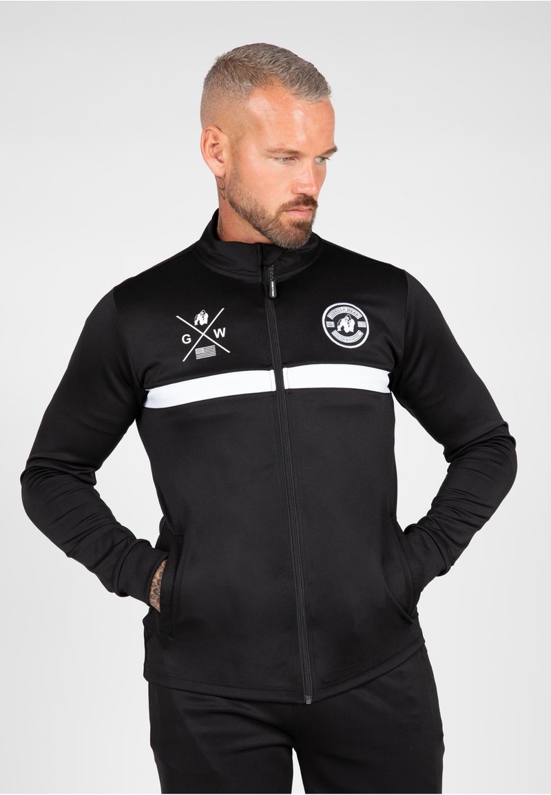 Zwarte zip-up jas van gladde stof, met een witte horizontale streep en logo's op de borst. Heeft een hoge kraag en zijzakken.
