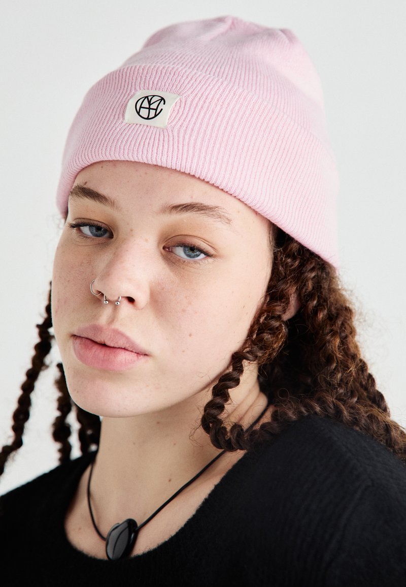 Pink knitted beanie with a ribbed texture, featuring a white label with a black logo. The background is light, emphasizing the hat's color.