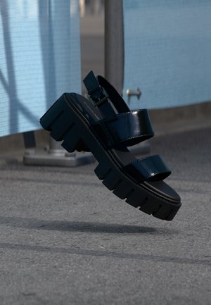 Svart chunky platåsandal med to stropper og spenne, svever i luften over grå fortau og blått stoffbakgrunn.