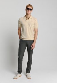 Beige gestippeld polo-shirt met een kraag, gecombineerd met donkergrijze slim-fit broek en lichtgekleurde sneakers. Accessoires omvatten een zonnebril en een sjaal.