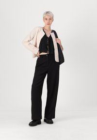 Calças largas pretas, cardigan rosa cropped e blusa preta de renda com bordas recortadas. Modelo segura uma bolsa preta acolchoada.