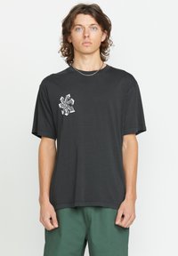 Zwarte katoenen t-shirt met korte mouwen, voorzien van een grafisch ontwerp op de linkerborst, gecombineerd met groene shorts, op een neutrale achtergrond.