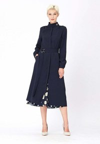 Manteau long marine avec un col haut, taille ceinturée et accents de boutons. En dessous, une robe à motif floral. Texture lisse.