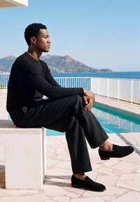 Homme en pull noir, pantalon et chaussures assis sur un banc blanc près de la piscine, avec vue sur la mer et les montagnes en plein jour clair.