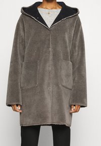 Cappotto di peluche grigio con un ampio cappuccio, due tasche frontali e un colletto scuro a contrasto. Tessuto morbido e chiusura con bottoni.