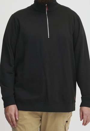 Homme portant un pull noir à manches longues avec un col zippé partiel et un pantalon beige, debout devant un fond neutre.