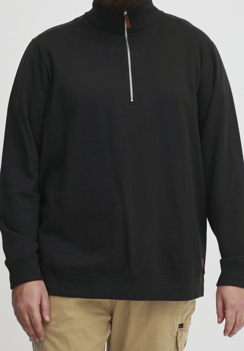 Homme portant un pull noir à manches longues avec un col zippé partiel et un pantalon beige, debout devant un fond neutre.