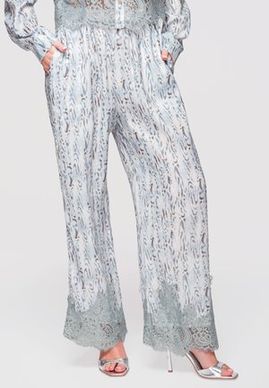 Pantaloni larghi con motivo astratto blu e marrone, orli in pizzo verde, indossati con sandali argento con tacco alto e punta aperta, mani in tasca.