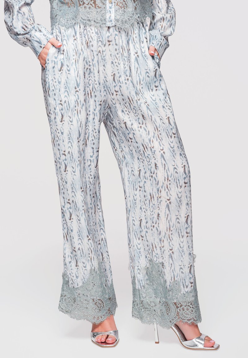 Pantalon ample à motif abstrait bleu et marron, ourlets en dentelle verte, porté avec des sandales argentées à talons hauts et bouts ouverts, mains dans les poches.
