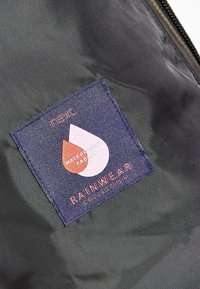 Gros plan d'une étiquette en tissu imperméable cousue à l'intérieur d'un vêtement de pluie gris foncé, avec le nom de la marque "next" et des icônes de gouttes d'eau.