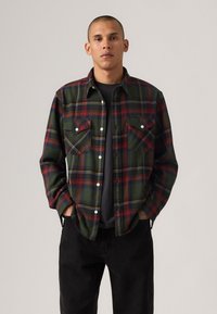 Green and red plaid flannel shirt with two chest pockets, white buttons, and a relaxed fit, worn over a black shirt, paired with black pants.