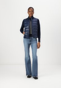 Gilet imbottito blu navy con colletto alto sopra una maglietta a righe, abbinato a jeans a zampa d'elefante blu e scarpe nere. Tessuto morbido, vestibilità casual.