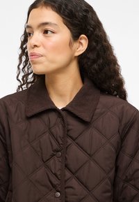 Jovem mulher com cabelo encaracolado escuro, vestindo uma jaqueta acolchoada marrom com um colarinho de veludo, olhando para o lado contra um fundo branco.