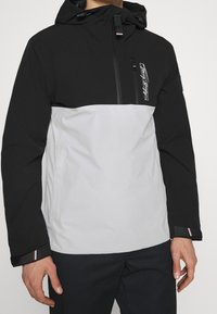 Nepromokavá bunda s černou horní částí a šedou dolní částí, s kapsou na zip na přední straně, nastavitelnou kapucí a barevnými akcenty na manžetách.