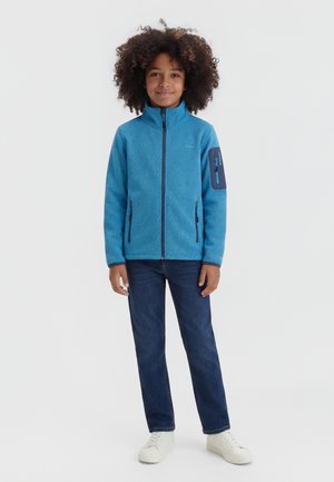 Παιδί με κατσαρά μαλλιά, φορώντας μπλε φλις μπουφάν με φερμουάρ, σκούρα τζιν και λευκά αθλητικά παπούτσια, στέκεται μπροστά σε φόντο απλό.