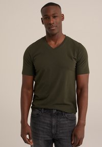 Camiseta de manga corta en verde oscuro con cuello en V, hecha de tela suave. Combinada con jeans negros desgastados, ajustados al cuerpo. Diseño y textura simples.