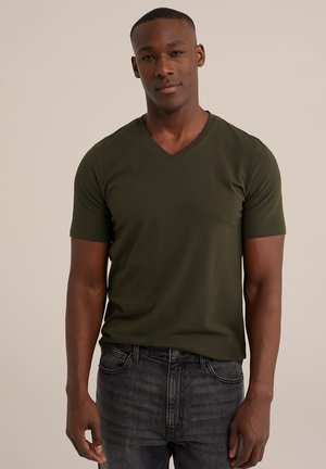 Camiseta de manga corta en verde oscuro con cuello en V, hecha de tela suave. Combinada con jeans negros desgastados, ajustados al cuerpo. Diseño y textura simples.