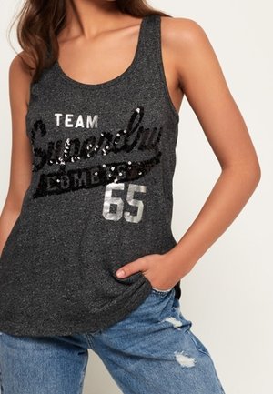 Bezrekawnik w ciemnoszarym kolorze z czarnym tekstem z cekinów "Superdry" i srebrnym napisem "Team 65", noszony z podartymi niebieskimi dżinsami, jedna ręka w kieszeni.
