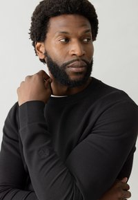 Felpa nera con polsini a coste e un logo discreto sulla manica sinistra. Materiale morbido con un classico scollo tondo.