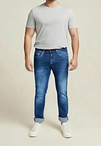 Mann trägt hellgraues kurzärmliges T-Shirt, blaue hochgekrempelte Jeans und weiße Sneakers, steht vor beigem Hintergrund.