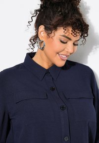 Camisa de color marino con cierre de botones, dos bolsillos en el pecho y cuello. Presenta una tela texturizada y botones lisos. Se ven rizos en el cabello y pendientes de aro.