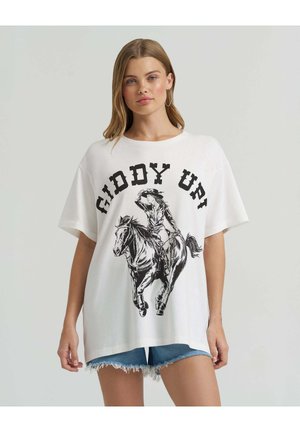 Frau trägt ein weißes, übergroßes T-Shirt mit schwarzem "GIDDY UP!"-Schriftzug und Grafik eines Cowboys auf einem Pferd, kombiniert mit ausgefransten Jeansshorts.