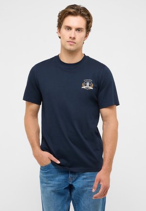 Young man with light brown hair wearing a navy blue t-shirt with a small logo on the chest and blue jeans, standing with one hand in pocket.