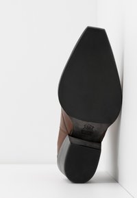 Podeszwa brązowego skórzanego buta eleganckiego z warstwowym obcasem, z widoczną czarną gumową podeszwą i wytłoczonym logotypem w pobliżu części łuku stopy.