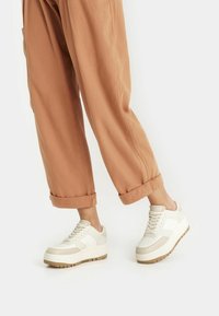 Platformové tenisky v off-white barvě s hladkým koženým povrchem, perforovanou špičkou a béžovými akcenty, spárované s ohrnutými hnědými kalhotami.