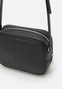 Černá malá crossbody taška Calvin Klein Jeans se stříbrným zipem a nastavitelným popruhem na bílém pozadí.