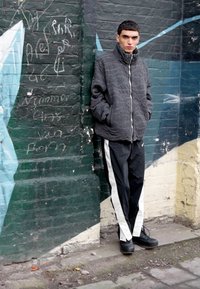 Giacca nera imbottita con colletto alto, abbinata a pantaloni neri con strisce bianche sui lati. Il modello si trova di fronte a un muro di mattoni con graffiti.