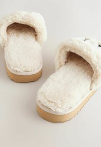Lynede, krammet cremefarvede sutsko med en blød tekstur, der har en tykk beige sål og rundet design. Inderforingen er fluffy.