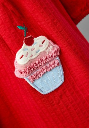 Roter Stoff mit einem Cupcake-Patch, der mit einer Kirsche obendrauf, pinker Glasur und blauen Cupcake-Boden verziert ist, akzentuiert mit Rüschen und dekorativen Nähten.