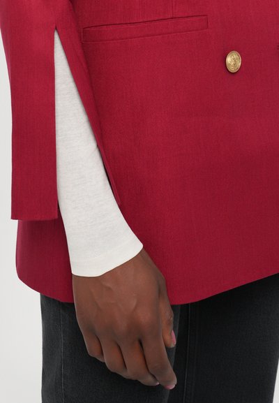 Red tailored blazer with a structured fit, single gold button, and side slits. White long-sleeve top underneath. Dark jeans visible.