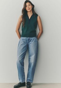 Marineblauw collared mouwloos topje, gecombineerd met lichtblauwe high-waisted denim jeans en zwarte instappers. Zachte textuur, comfortabele pasvorm en casual stijl.