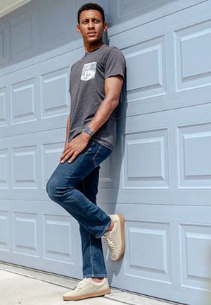 Junger Mann in grauem T-Shirt, blauen Jeans und weißen Sneakers lehnt an einer grauen Garagentür und schaut zur Seite mit entspannten Händen.