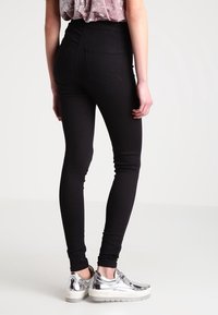 Jeans skinny de tiro alto en negro con bolsillos traseros, combinados con zapatillas de deporte plateadas, exhibiendo un diseño ajustado y una tela elástica.