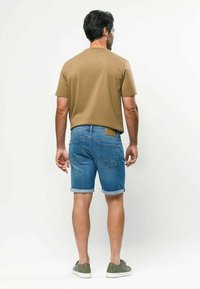 Ein Mann steht mit dem Rücken zur Kamera, trägt ein hellbraunes kurzärmeliges Hemd, blau hochgekrempelte Jeansshorts und olivgrüne Turnschuhe.