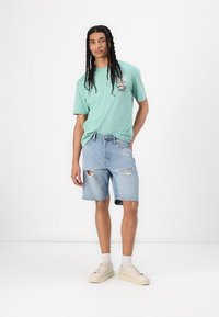 Tricou de culoare verde mentă cu un design grafic, asortat cu pantaloni scurți din denim albastru deschis, cu margini din franjuri și detalii uzate. Adidași crem cu sireturi.