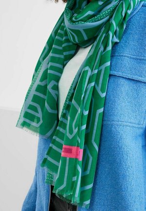 Écharpe verte avec des motifs géométriques bleu clair et un détail rectangulaire rose, drapée sur un manteau bleu texturé et un haut blanc.