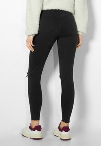 Jeans negros de talle alto con un diseño ajustado, que presentan áreas desgastadas en las rodillas, acentuados con un dobladillo deshilachado y combinados con zapatillas blancas gruesas.