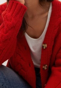 Cardigan en grosse maille rouge avec un motif en torsades texturées, orné de grands boutons dorés, porté par-dessus une chemise blanche, avec un jean bleu visible.