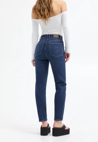 Jeans de tiro alto en azul con un diseño de pierna recta, que cuentan con bolsillos traseros y una etiqueta de cuero marrón en la cintura. Combinados con sandalias de plataforma negras.